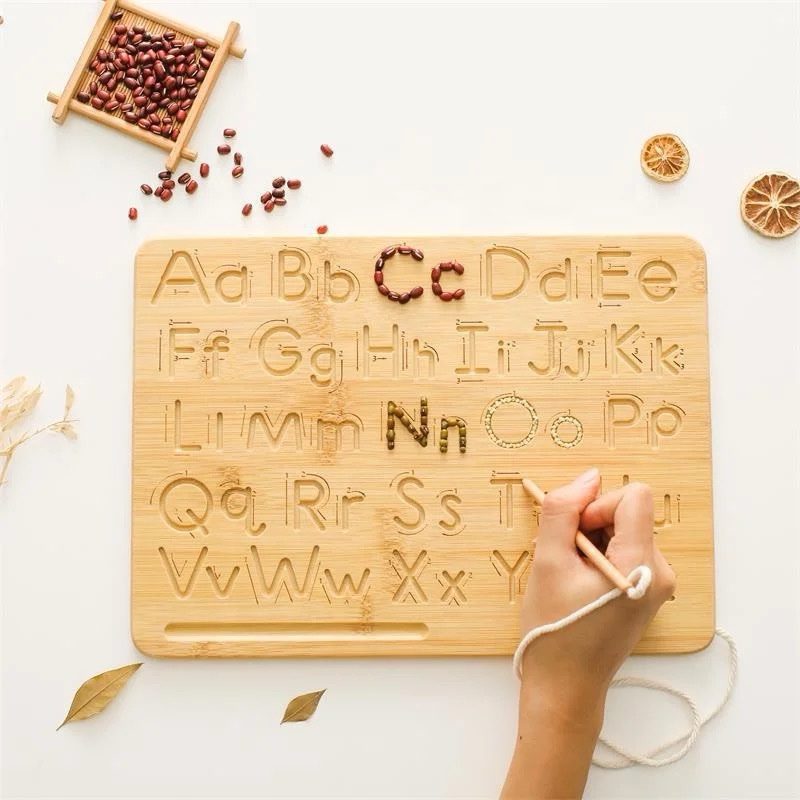 A wooden double-sided Montessori tracing board featuring engraved uppercase and lowercase alphabets, with a child using a wooden stylus to trace letters.