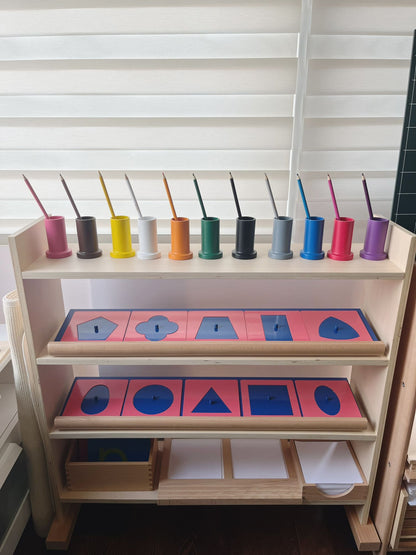 Montessori metal inset cabinet arranged with pencil holders, metal inset shapes, and paper boxes for writing activities.