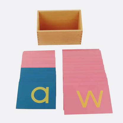 Spread of Montessori lowercase sandpaper letters in pink and blue cards placed beside an empty wooden storage box.