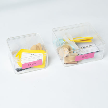 Two transparent storage boxes containing Montessori singular and plural word cards with small objects and labels for organizing the language activity materials.