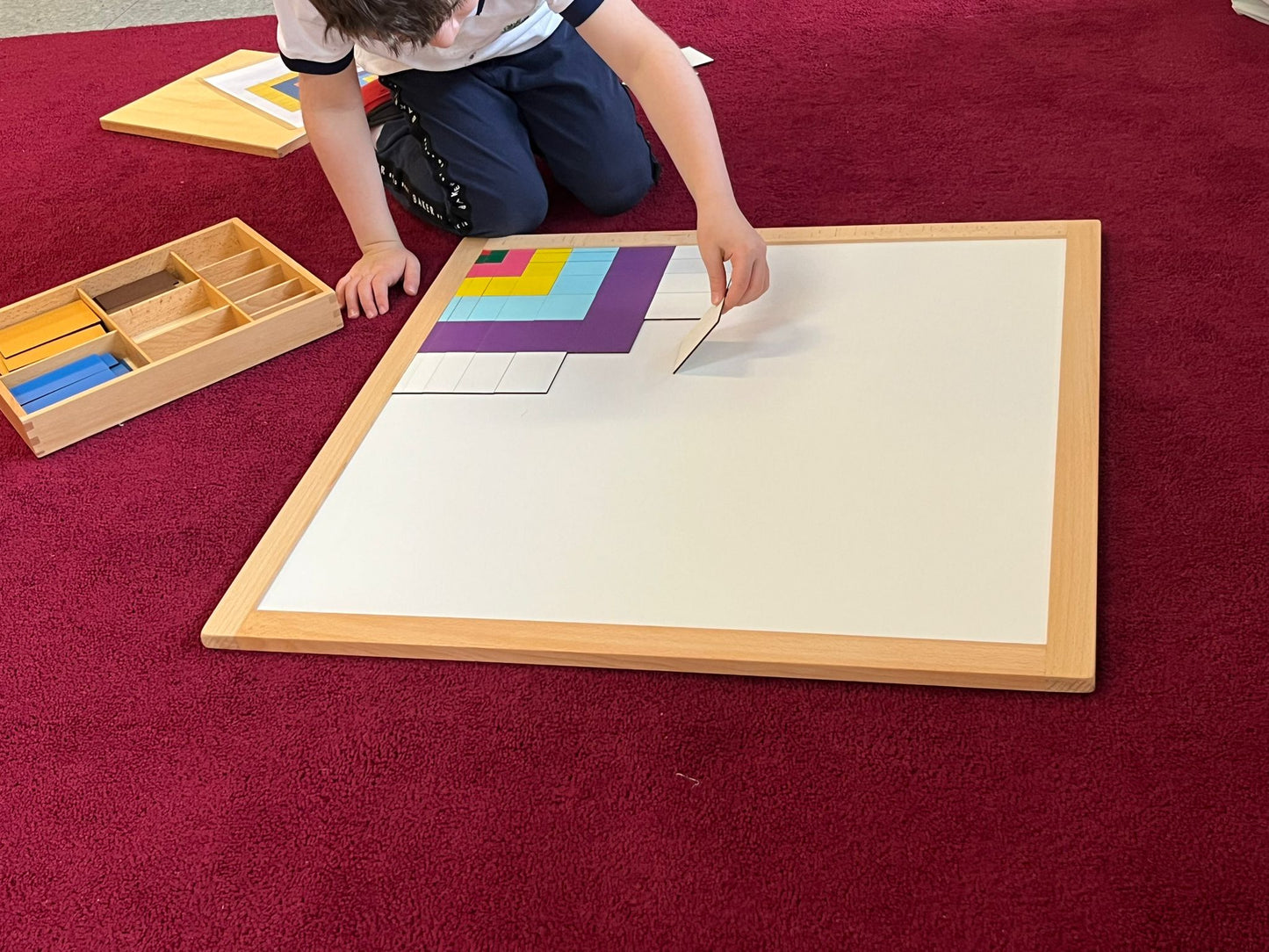 Child using a large Montessori wooden geometry board with colorful square insets on a red carpet.