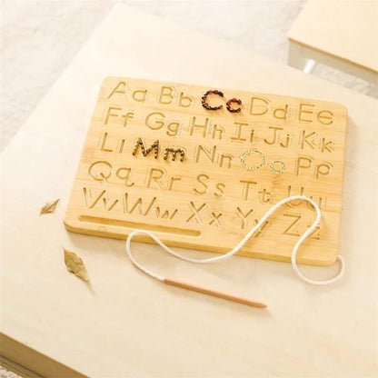 A wooden tracing board with engraved uppercase and lowercase letters placed on a table, with a child’s tracing stylus positioned beside it.