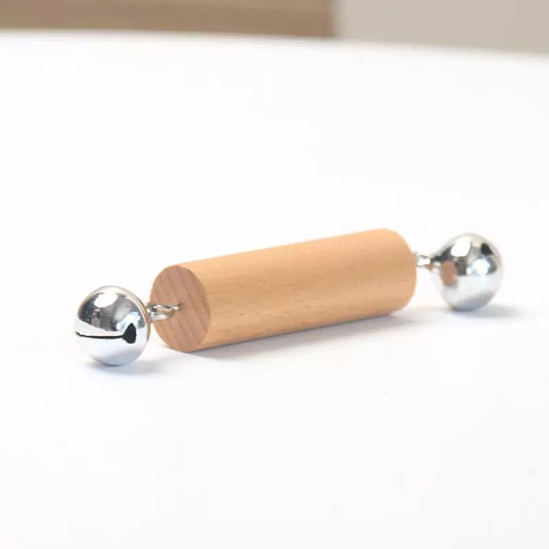 Wooden Montessori rattle toy with a smooth cylindrical wooden handle and silver metal bells attached on both ends, placed on a light background.